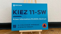 Das Bild zeigt ein blaues Schild der Stadt Bochum, auf dem "Hinweisschild zum Krisen-Informations-Ersthilfe-Zentrum", sowie die Adresse und die genauen Längen- und Breitengrade des Zentrums stehen.